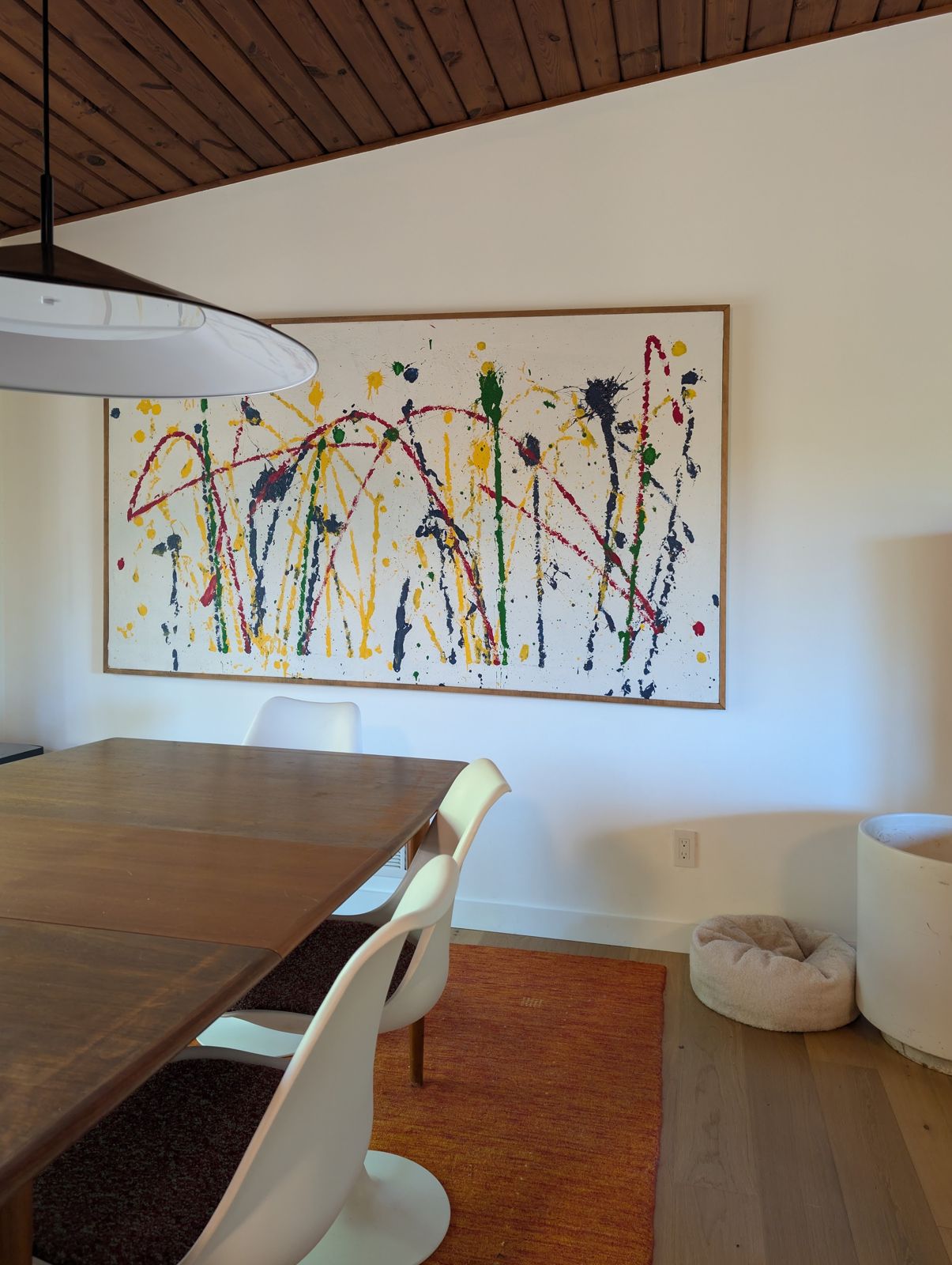 The dining room: a wood plank ceiling, a large drum pendant, a long wood table with white tulip chairs, an orange rug and a colorful drip painting