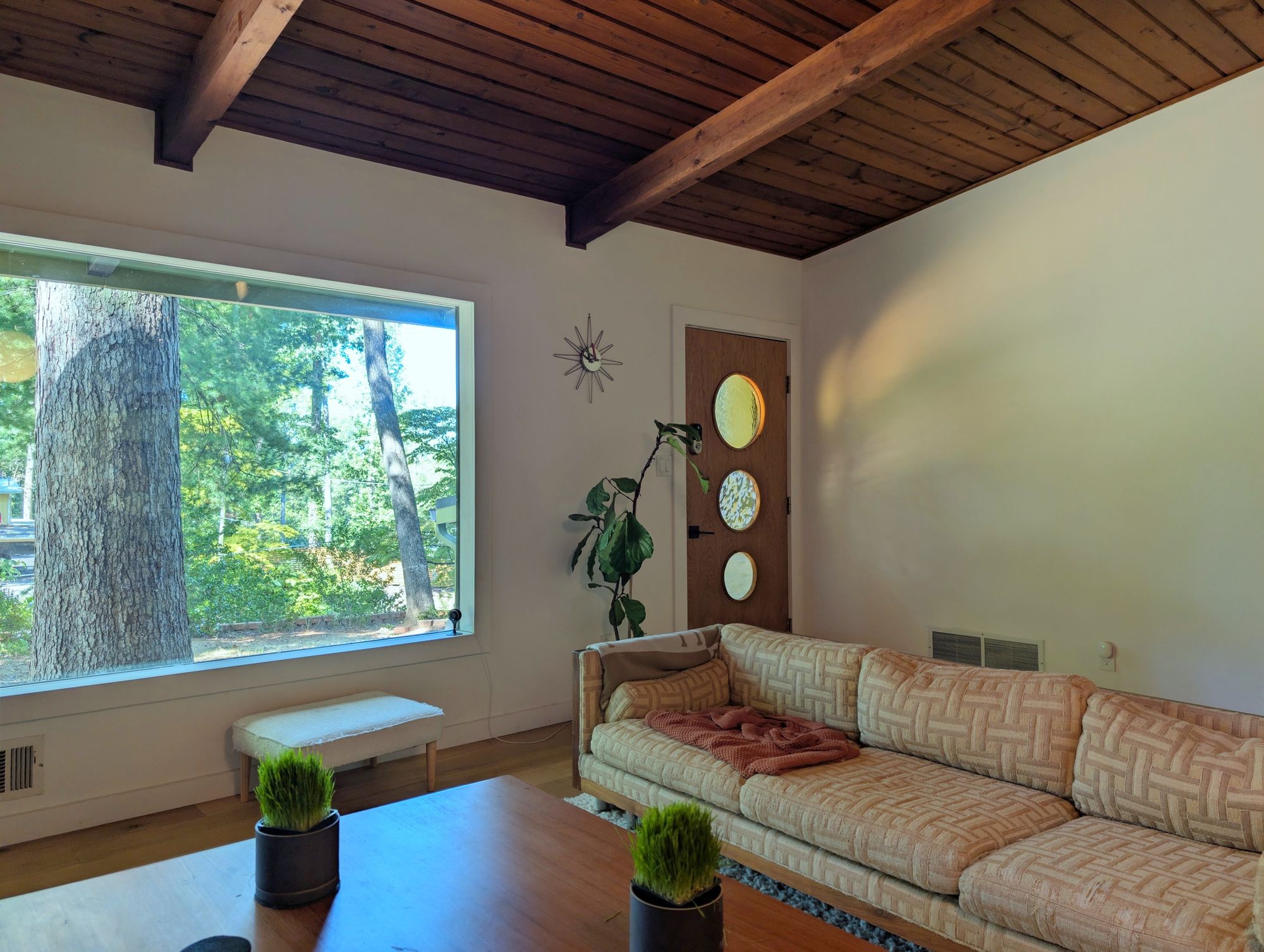 The family room: a wide picture window onto tall trees, a wood plank ceiling on beams, a sunburst clock and a basket weave sofa
