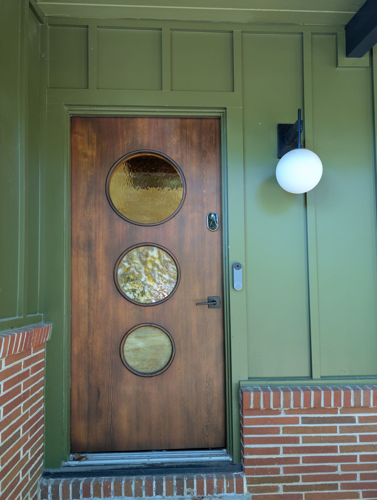 The wood front door with three round porthole windows, a globe sconce and a smart lock, set in olive green board and batten siding above a brick base